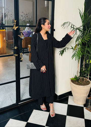 A woman standing in a stylish interior setting, next to a potted plant, wearing a black outfit with a V-neck top and midi skirt.